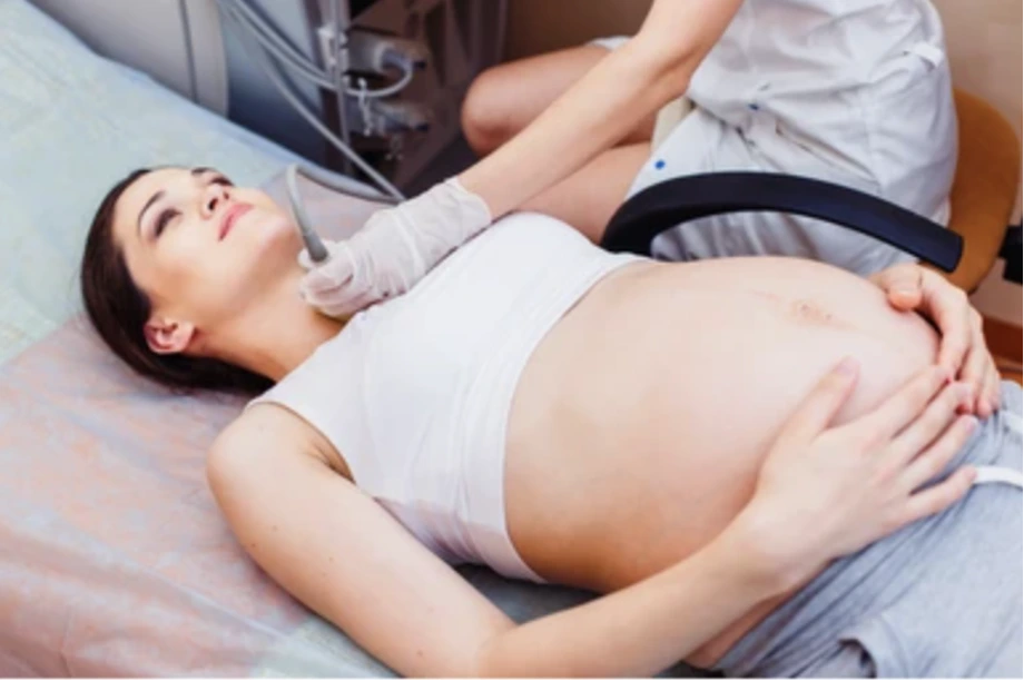 Preg Picture 2 - Dr Sandeep Donagaon Pregnant woman lying on a hospital bed with hands resting on her baby bump as a clinician assists nearby.
