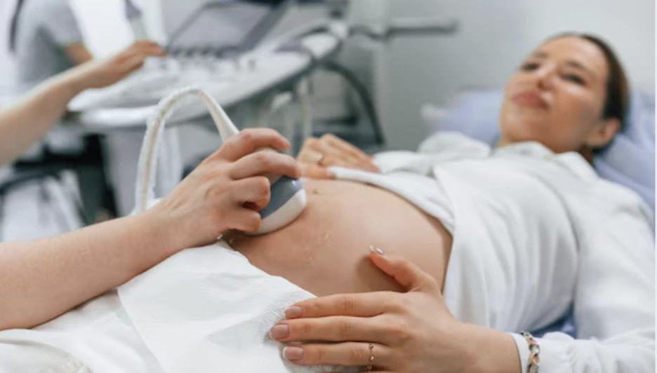Preg Picture 3 - Dr Sandeep Donagaon Pregnant woman lying on a medical bed while a clinician uses an ultrasound probe on her abdomen. Slim device in the clinician's hand shows an obstetric ultrasound in progress.