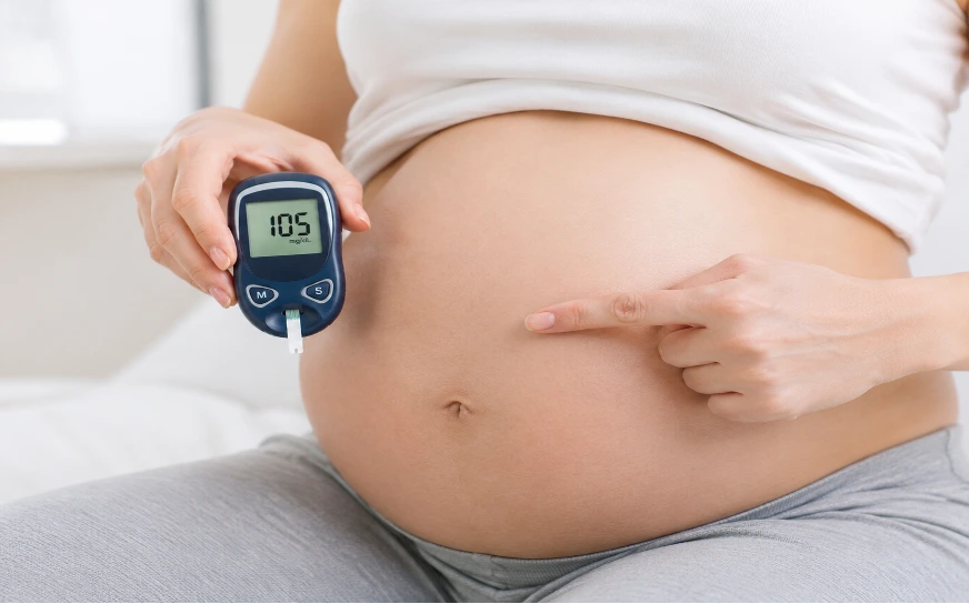 Pregnant woman sits with a glucose meter showing 105 mg/dL while pointing to her bare belly to indicate gestational diabetes monitoring.