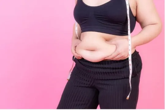 Person in a black sports bra and pants measures their waist with a tape measure against a pink background, focusing on midsection.