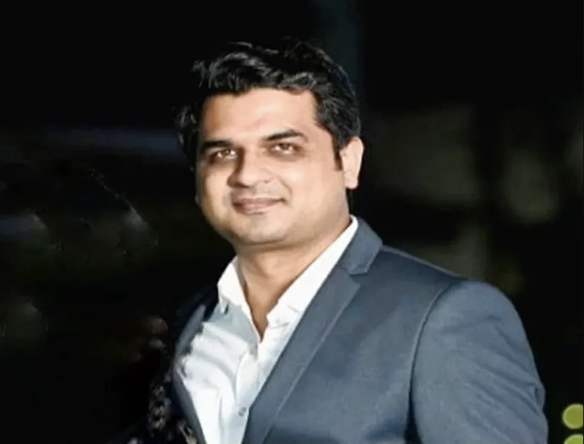 Man in a light gray suit and white shirt smiles at the camera against a dark background.